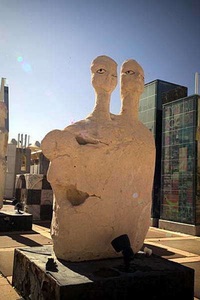Escultura de dos cabezas sobre una base oscura al aire libre, ante edificios acristalados.