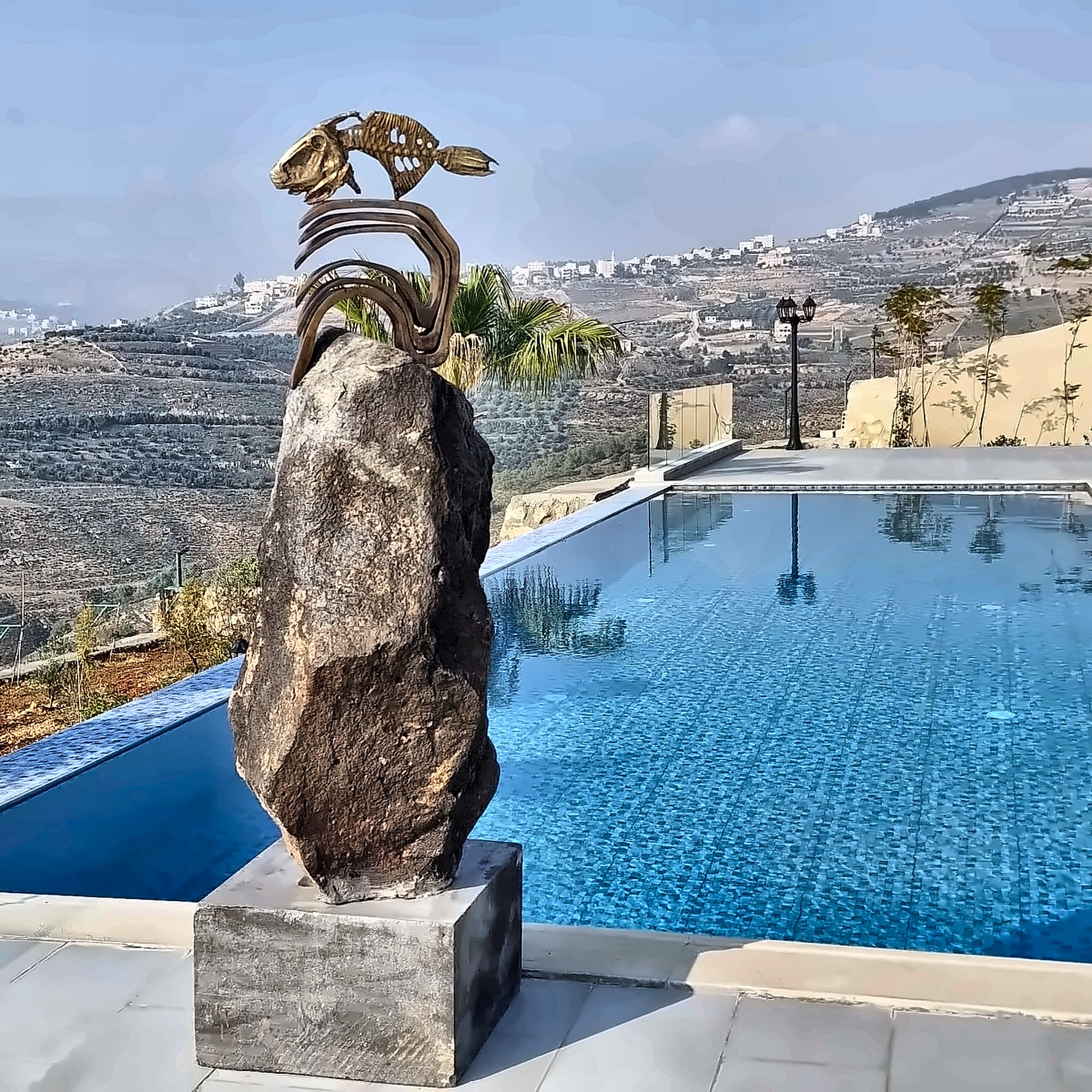 A rough basalt boulder on a black plinth beside an infinity pool, carrying a gilded bronze fish skeleton and a wave form, with terraced hills beyond.