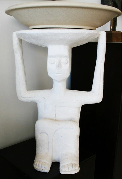 Una figura de yeso blanco con ambos brazos levantados, que sostiene un cuenco ancho sobre la cabeza.