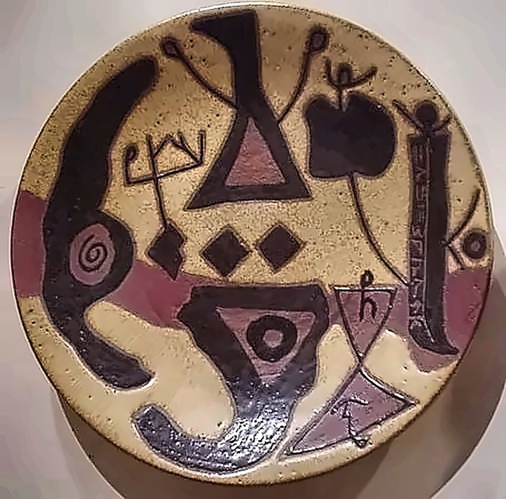 Plato de gres con formas negras pesadas, un triángulo, una espiral y una columna de escritura incisa a un lado.