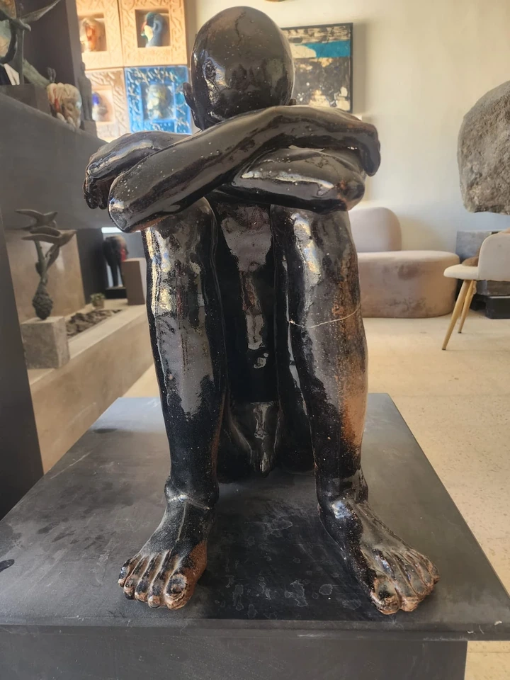 Vista frontal de la figura sentada de gres, con los pies apoyados y los brazos cruzados sobre las rodillas.