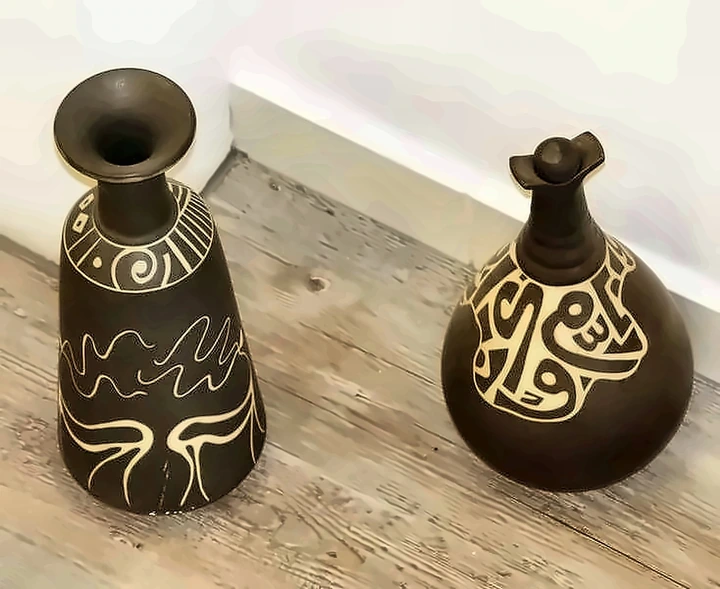 Dos vasijas oscuras, una cónica y otra redonda, atravesadas por línea y escritura en esgrafiado blanco.