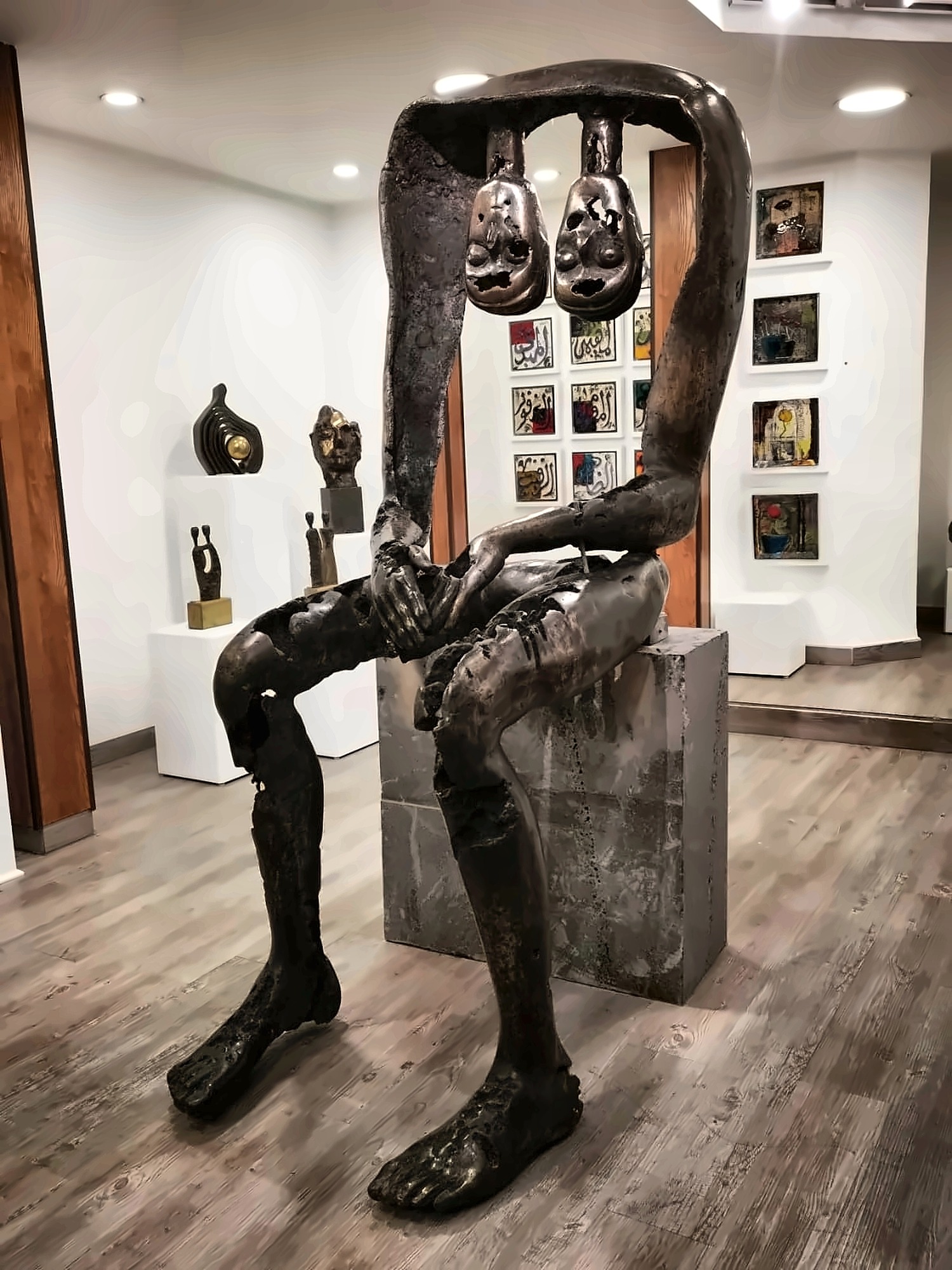 A monumental seated bronze figure in a gallery, its torso an open arch with two small pierced heads hanging inside it, legs at full length, hands folded over one knee.